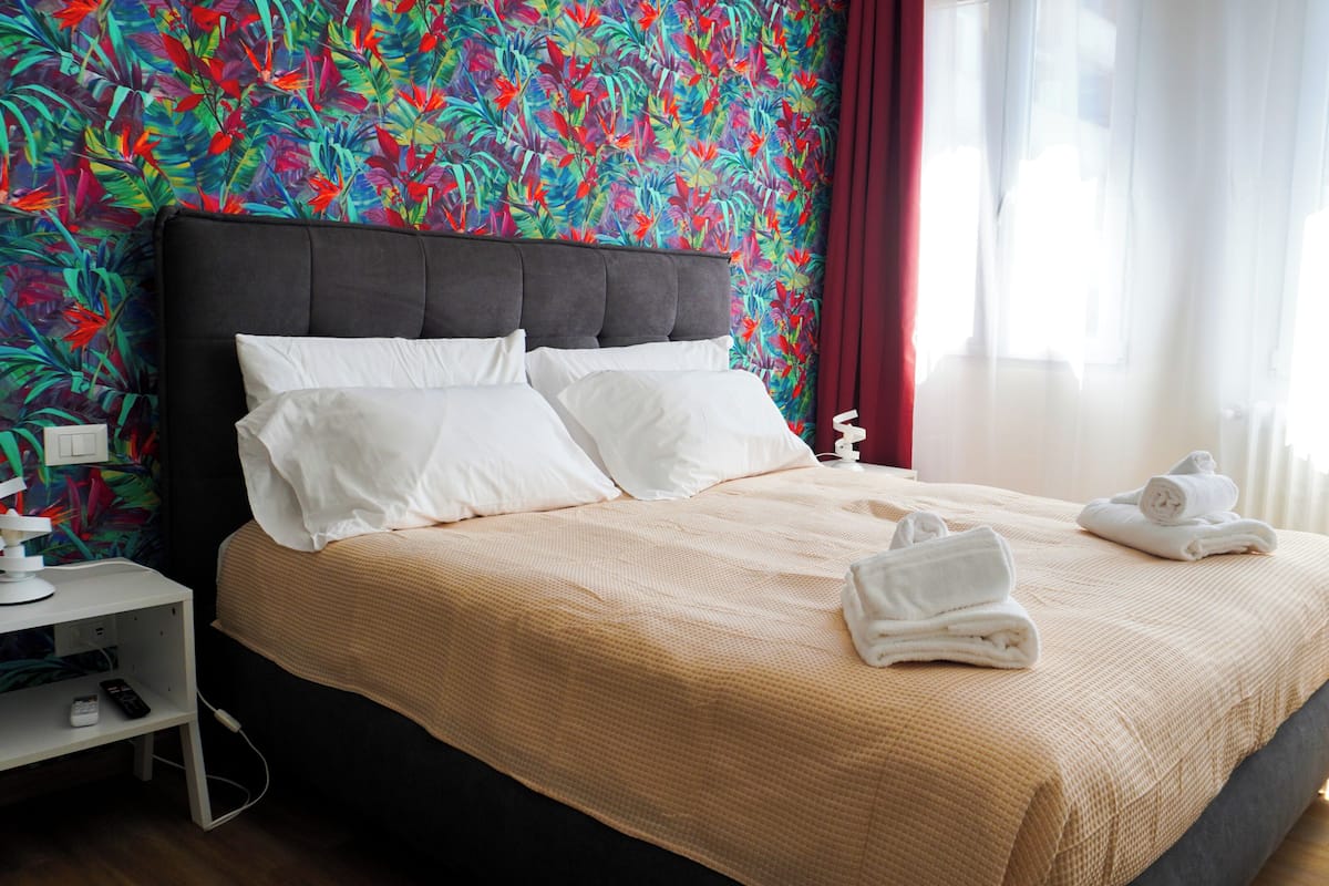 A hotel room with a bed, white pillows, a nightstand with a lamp, and a red curtain.