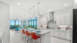 A modern kitchen with white cabinets, a large island with bar stools, and a view of the ocean through large windows. - Sunset Shores | Beachfront Condo w/ Premium Master, PVT Balcony & Upscale Decor (Melbourne Beach)
