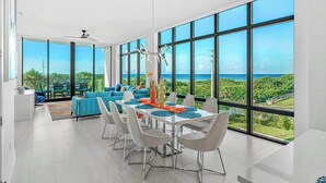 A modern dining area with a table set for six, large windows offering a view of the ocean, and a bright, airy room with a ceiling fan. - Sunset Shores | Beachfront Condo w/ Premium Master, PVT Balcony & Upscale Decor (Melbourne Beach)