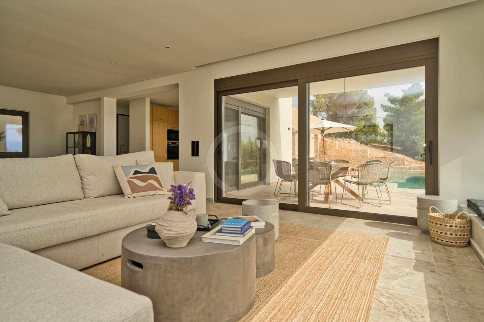 Ein modernes Wohnzimmer mit einer großen Couch, einem Couchtisch mit Büchern und einer Vase, und Blick auf eine Terrasse mit Stühlen und einem Pool.