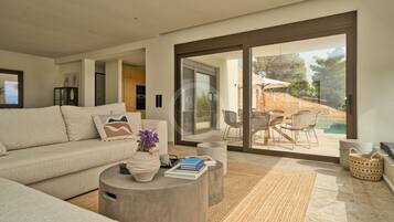 Ein modernes Wohnzimmer mit einer großen Couch, einem Couchtisch mit Büchern und einer Vase, und Blick auf eine Terrasse mit Stühlen und einem Pool.
