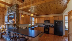 A spacious kitchen with wooden cabinets, a large island with bar stools, a stone accent wall, and a black refrigerator. - River House at Meadow Landing (Fleetwood)