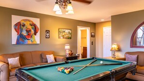 Een pooltafel in een kamer met een plafondventilator, een schilderij van een hond en een deur naar een andere kamer.