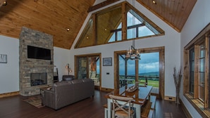 A spacious living room with a stone fireplace, a large sofa, and a dining area with a view of the outdoors. - Peregrine's Perch at Eagles Nest (Elk Park)
