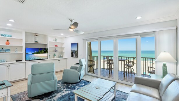 A spacious living room with a large TV, comfortable seating, and a balcony with a view of the beach.
