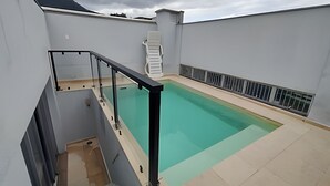 Una piscina en la azotea con barandilla de vidrio, una silla de descanso blanca y una escalera que conduce a un nivel superior.