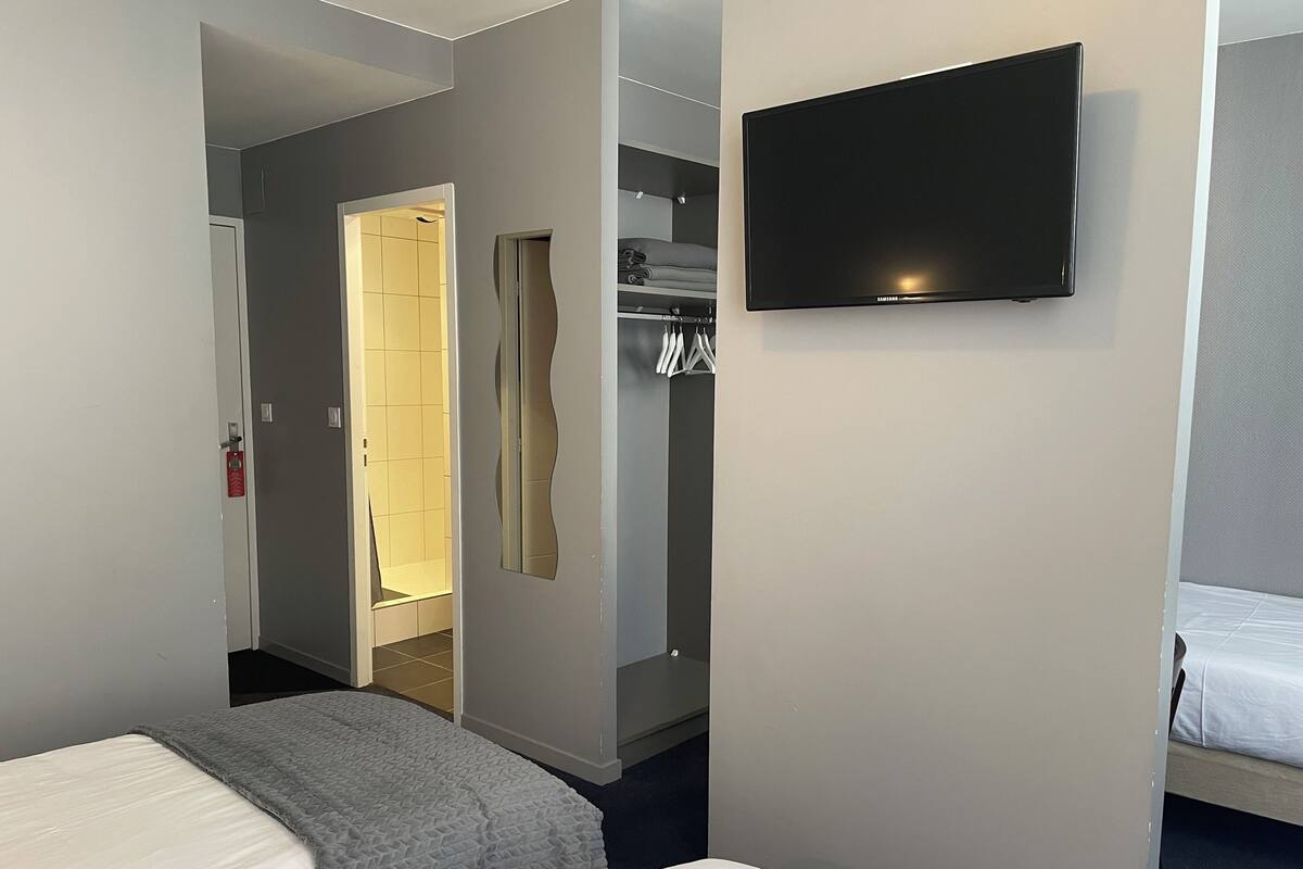 Una habitación de hotel con dos camas, un televisor montado en la pared, un armario con ropa colgada y un baño visible a través de una puerta abierta.