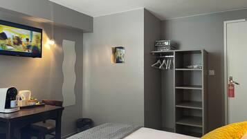 A hotel room with a bed, a desk with a coffee machine, a TV mounted on the wall, a wardrobe, and a shelf with hangers.
