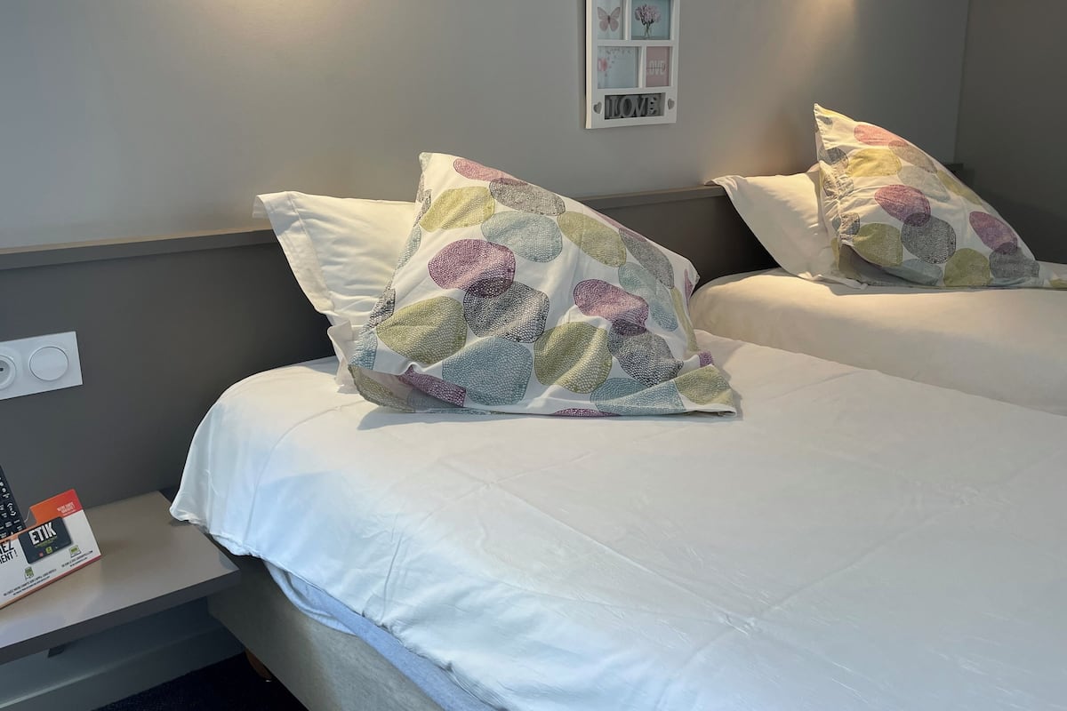 Habitación de hotel con dos camas, cada una con cubrecama blanco y almohada con estampado de hojas coloridas. Hay un cuadro colgado en la pared con una mariposa y flores, y un letrero que dice 'LOVE'.