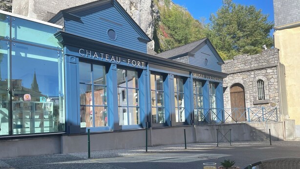 A cafe with a blue exterior, a stone building in the background, and a table with croissants, a glass of orange juice, and a teapot. - Castel de Mirambel (Lourdes)
