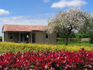 A two-story house with a red-tiled roof, a front porch, and a garden with red foliage and a flowering tree. - Parc Résidentiel de Vacances Arboré 9 Gites Avec Piscine Couverte et Chauffée (Le Ham)