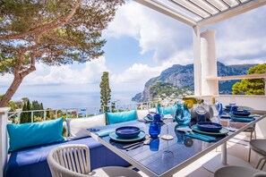 A dining table set with blue and white tableware, overlooking a coastal landscape with mountains and the sea. - Blue Essence (Capri)