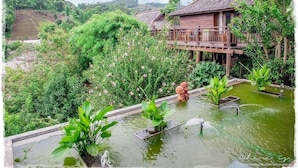 A traditional wooden house with a balcony, surrounded by lush greenery and a small pond with plants. - Baan Mon Muan  (Mae Rim)