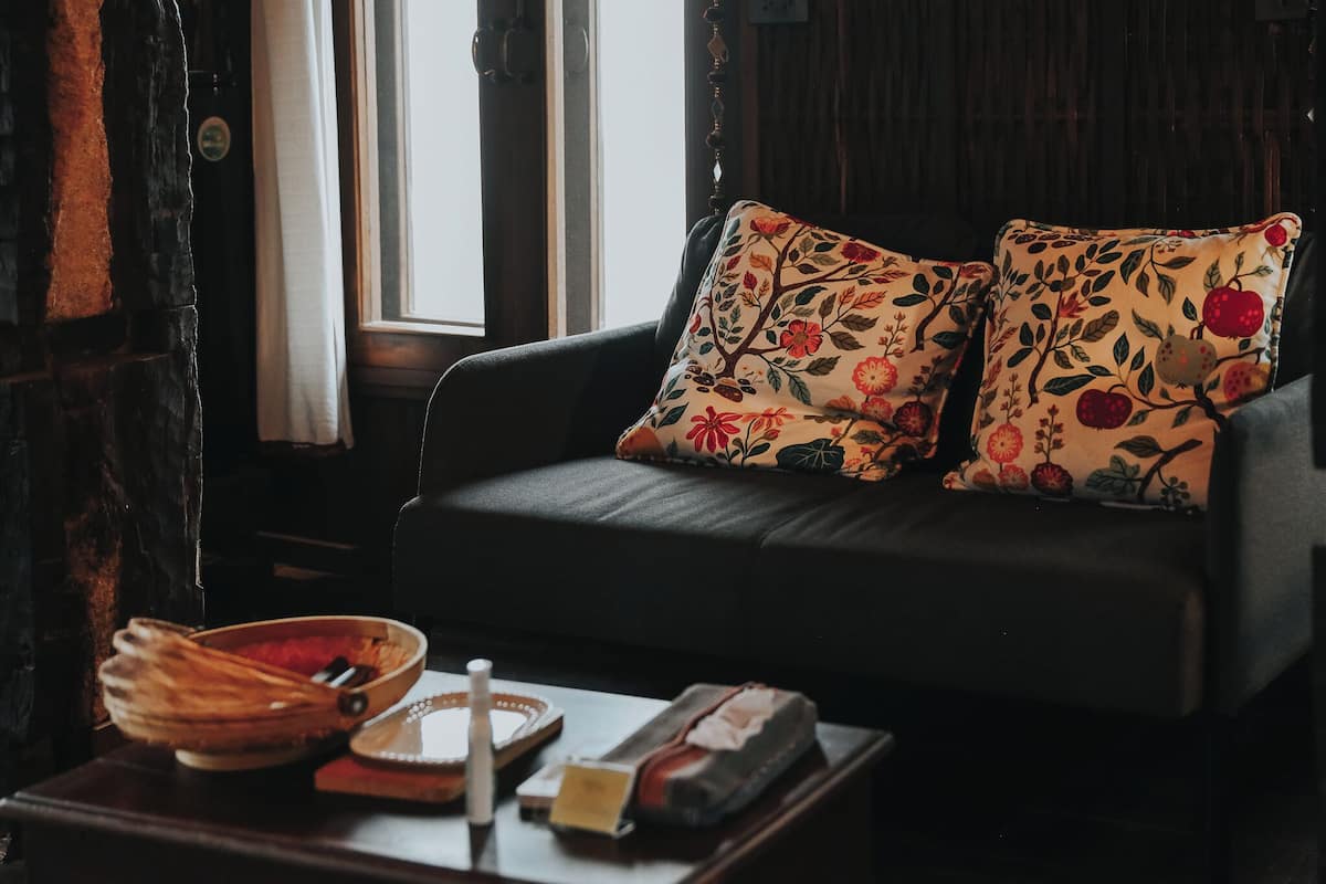 Un sofá de color oscuro con cojines de estampado floral, una mesita de madera con un cuenco, una vela y un libro.