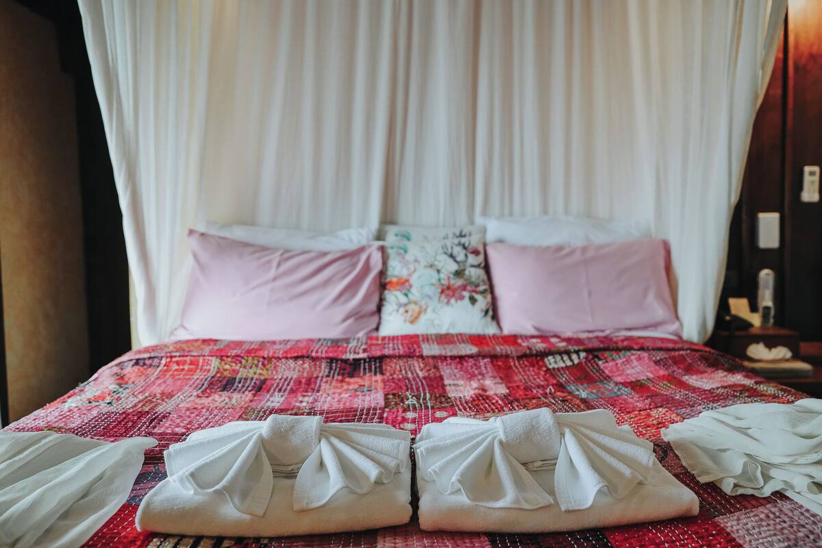 Una cama con una colcha de estampado floral, dos almohadas y toallas dobladas ordenadamente a un lado.
