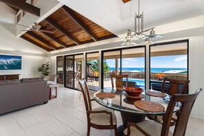 A spacious living area with a glass dining table, wooden chairs, a sofa, and a large window offering a view of the ocean. - Luxury 3BR Kona Dreams w/ Ocean Views, Private Pool, Central AC & Prime Location (Kailua Kona)