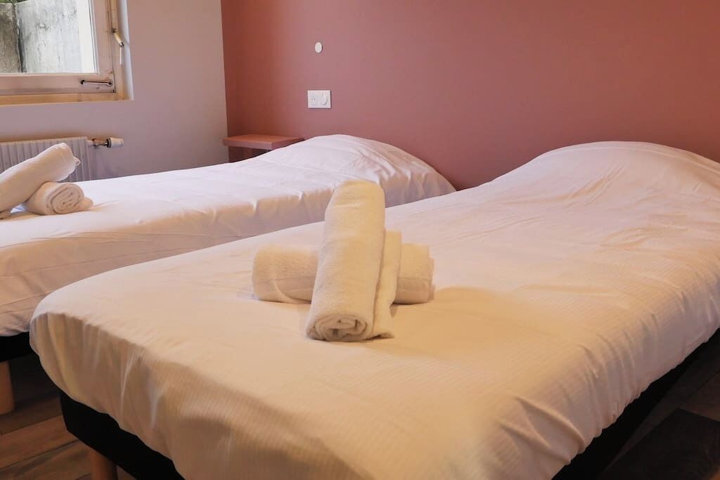 Two single beds with white linens and rolled towels, a window with a view, and a wall socket.