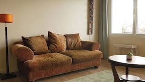 A living room with a brown sofa, a small round table with a plant, a floor lamp, and a window with curtains.