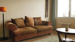 Een woonkamer met een bruine bank, een ronde tafel met een plant erop, een vloerlamp en een raam met gordijnen.