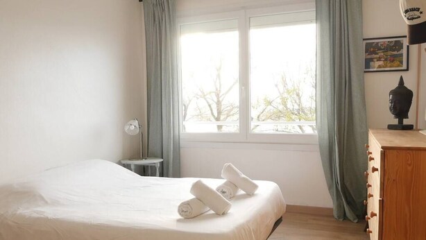 A bedroom with a bed, a small table, a lamp, a dresser, and a window with curtains.