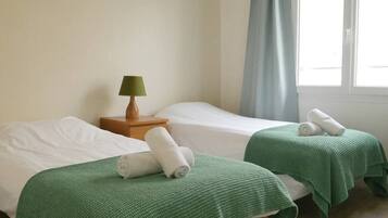 Ein Zimmer mit zwei Betten, jedes mit einer grünen Decke und aufgerollten Handtüchern. Zwischen den Betten steht ein Nachttisch mit einer Lampe.