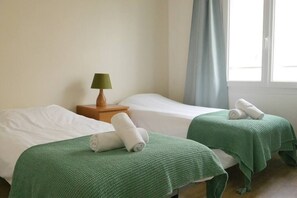 A room with two beds, each with a green blanket and rolled towels. There is a wooden nightstand with a lamp between the beds.