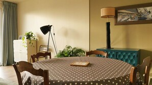 A dining area with a round table covered in a polka dot tablecloth, surrounded by wooden chairs. There is a lamp on a stand, a framed picture on the wall, and a small cabinet with a plant on top.
