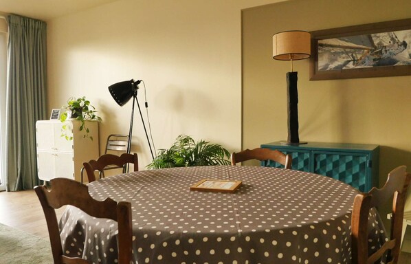 Un comedor con una mesa redonda cubierta por un mantel de lunares, rodeado de sillas de madera. Hay una lámpara en un atril, un cuadro en la pared y un mueble bajo con una planta encima.