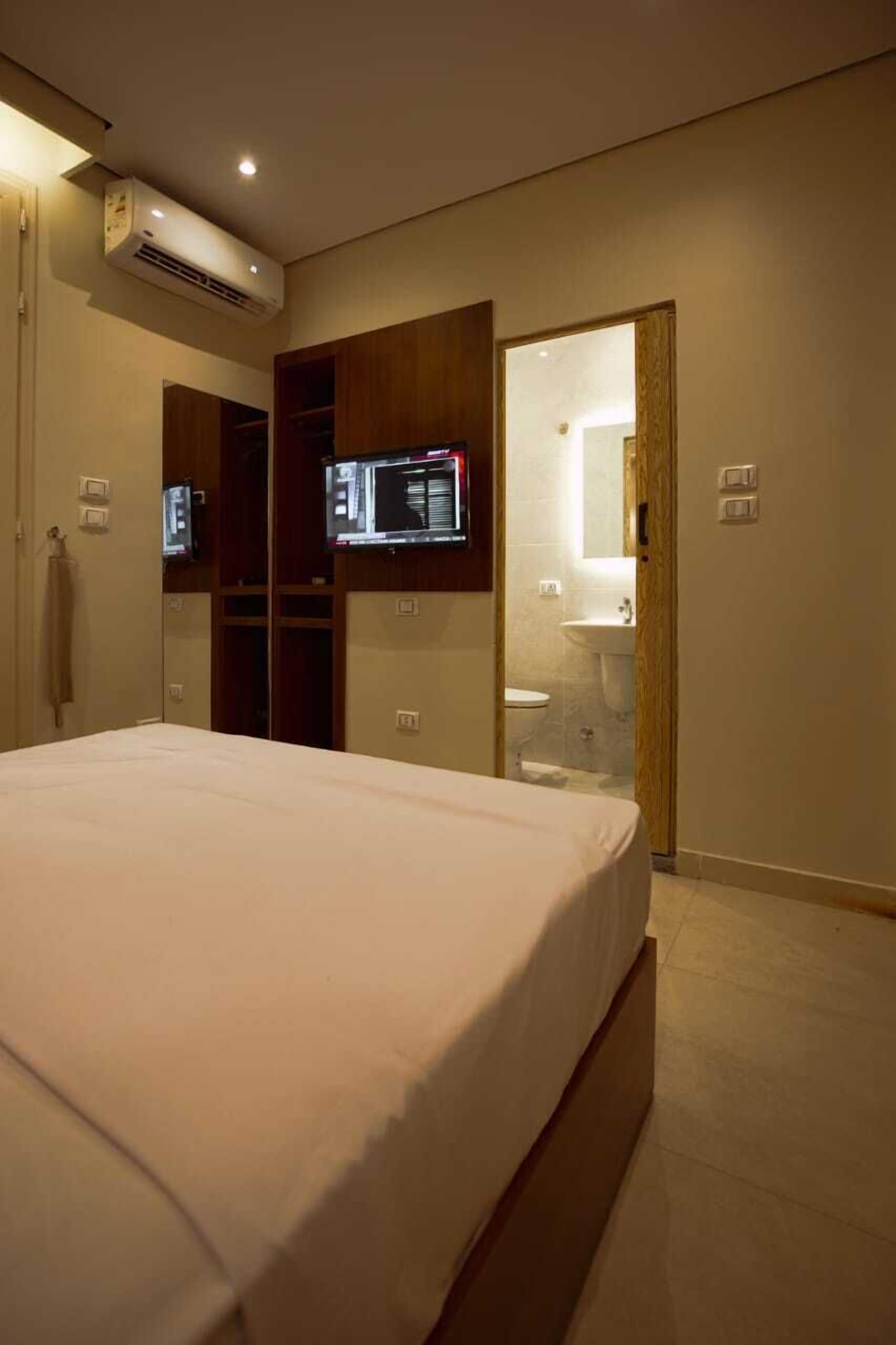 Un dormitorio con cama, unidad de aire acondicionado, televisor, espejo y baño visible a través de una puerta abierta.
