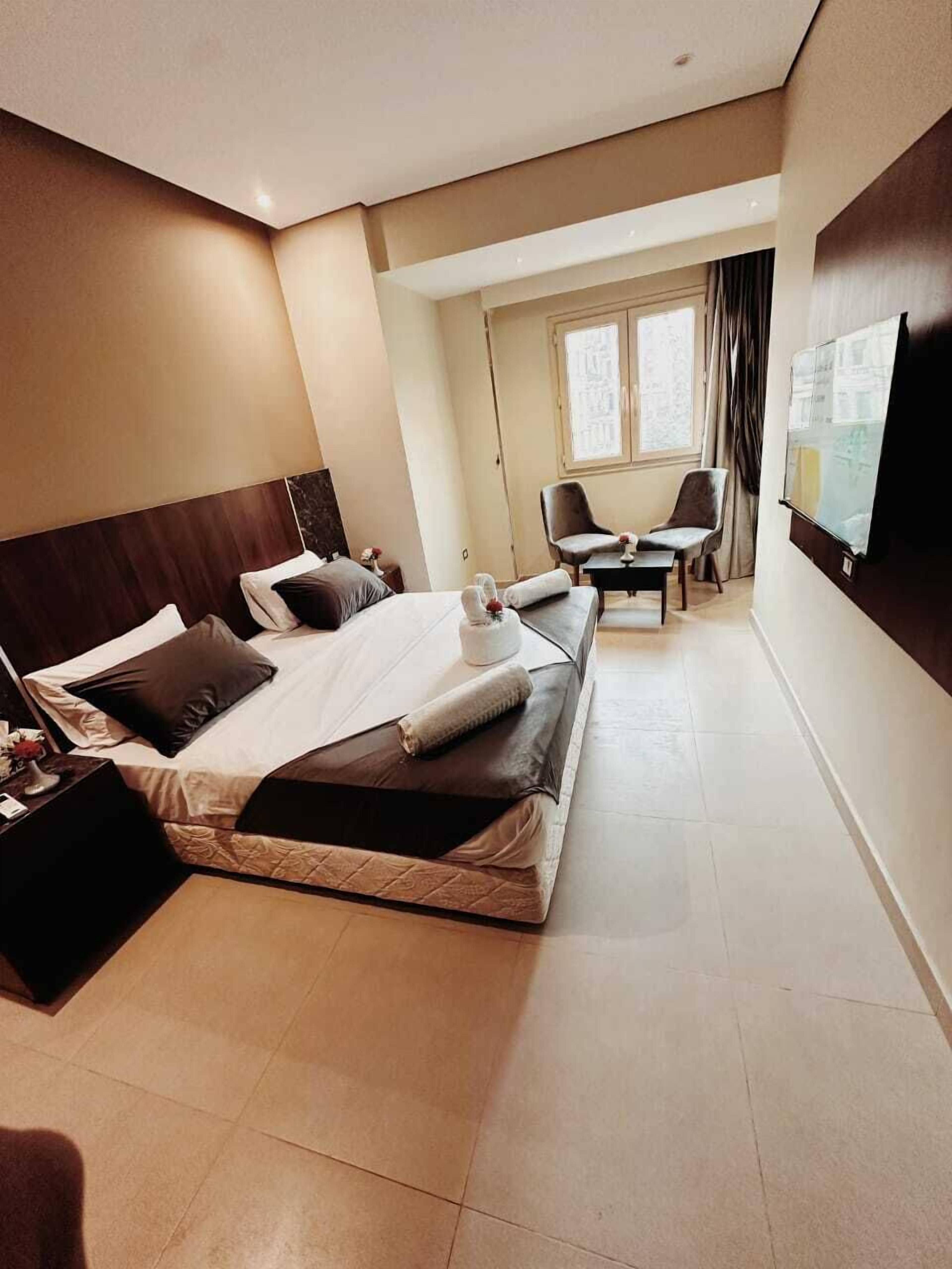 A hotel room with a large bed, a desk, two chairs, a TV, and a window with curtains.