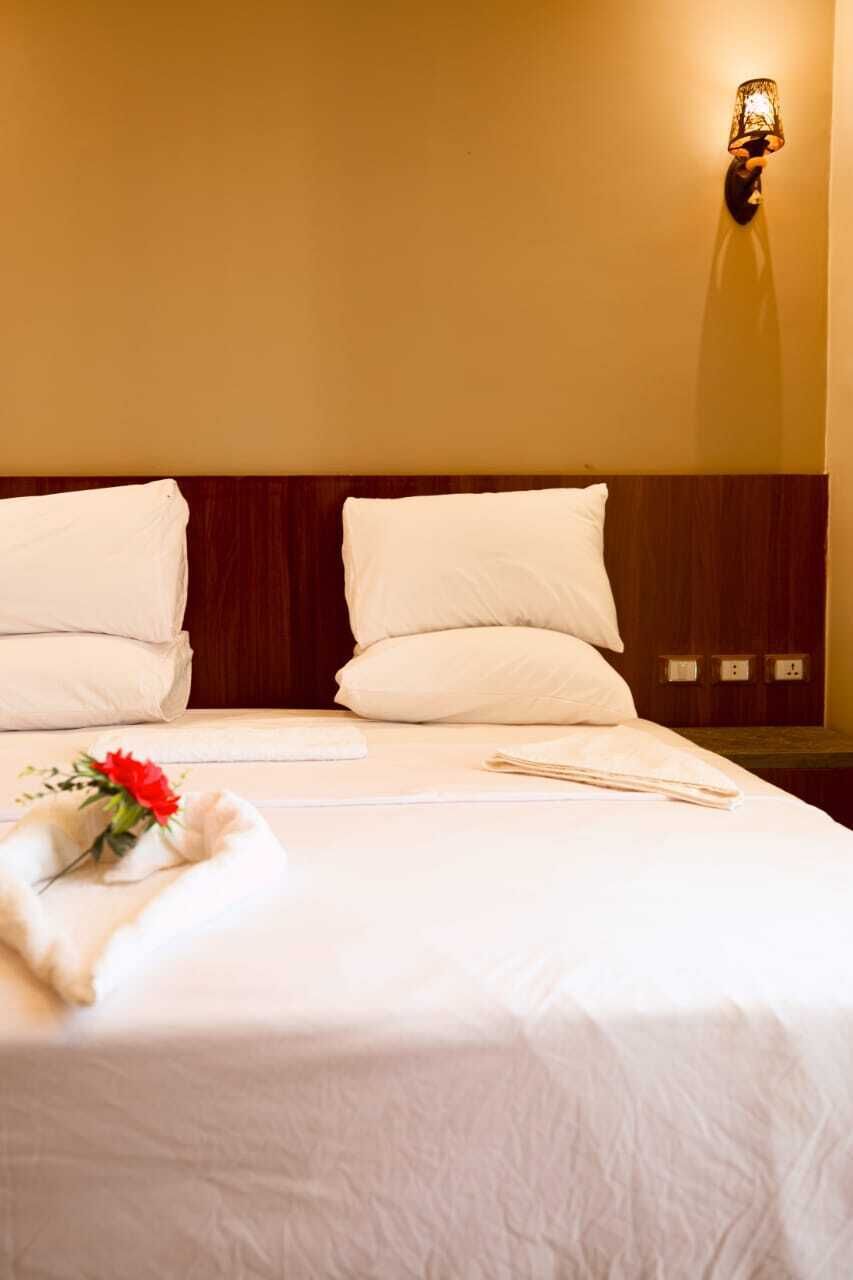 A neatly made bed with white linens and pillows, a bedside lamp, and a small red flower arrangement.