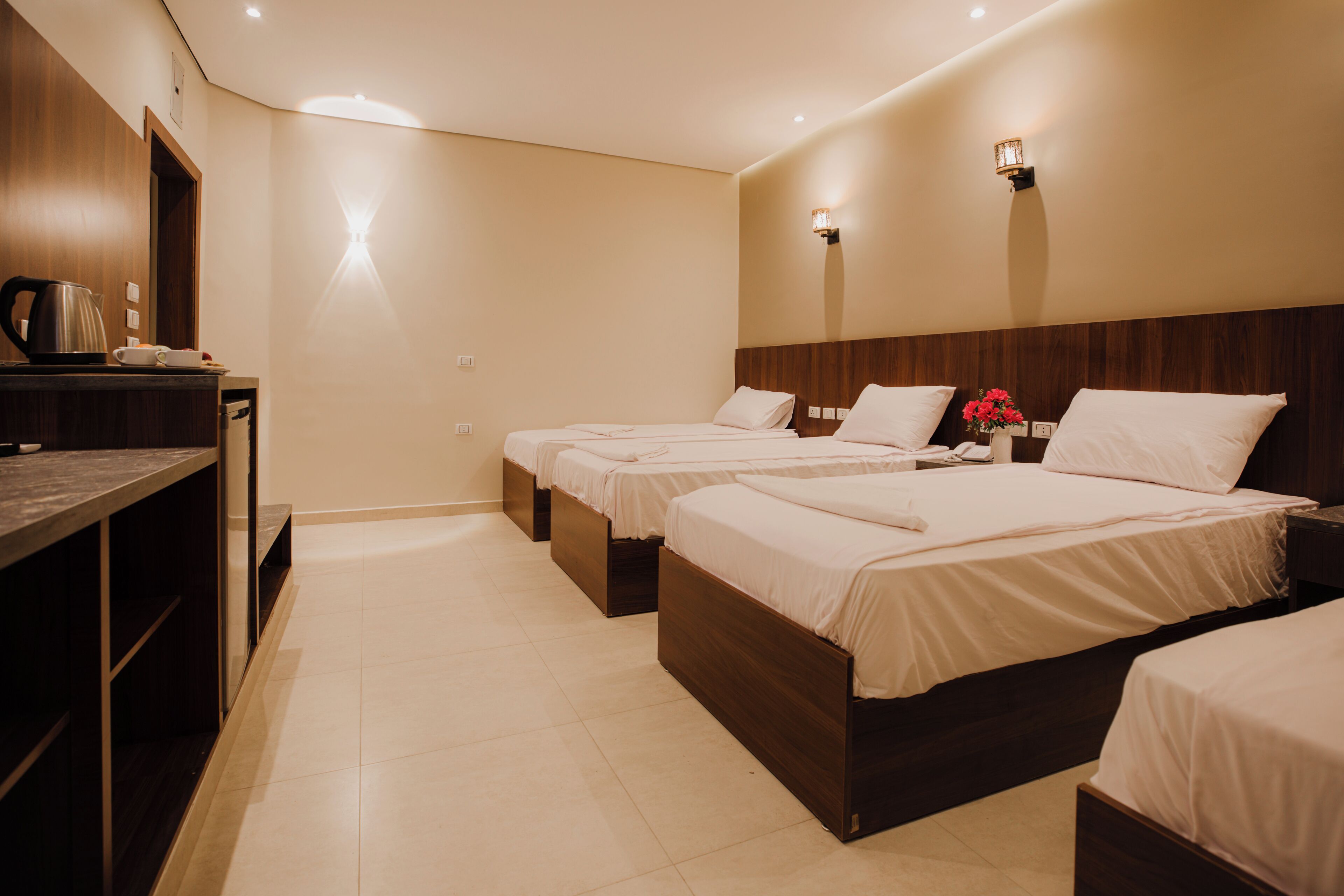 A hotel room with three single beds, a wooden headboard, a small table with a kettle, and a vase with flowers.