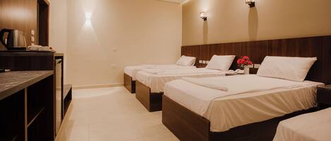 A hotel room with three single beds, a wooden headboard, a small table with a kettle, and a vase with flowers.