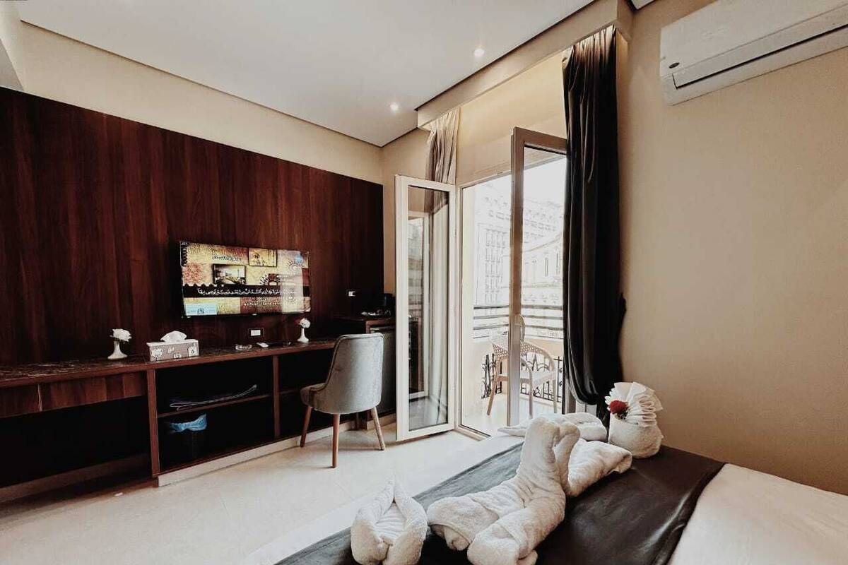 A modern hotel room with a large bed, a desk with a computer, a chair, and a balcony with a view.