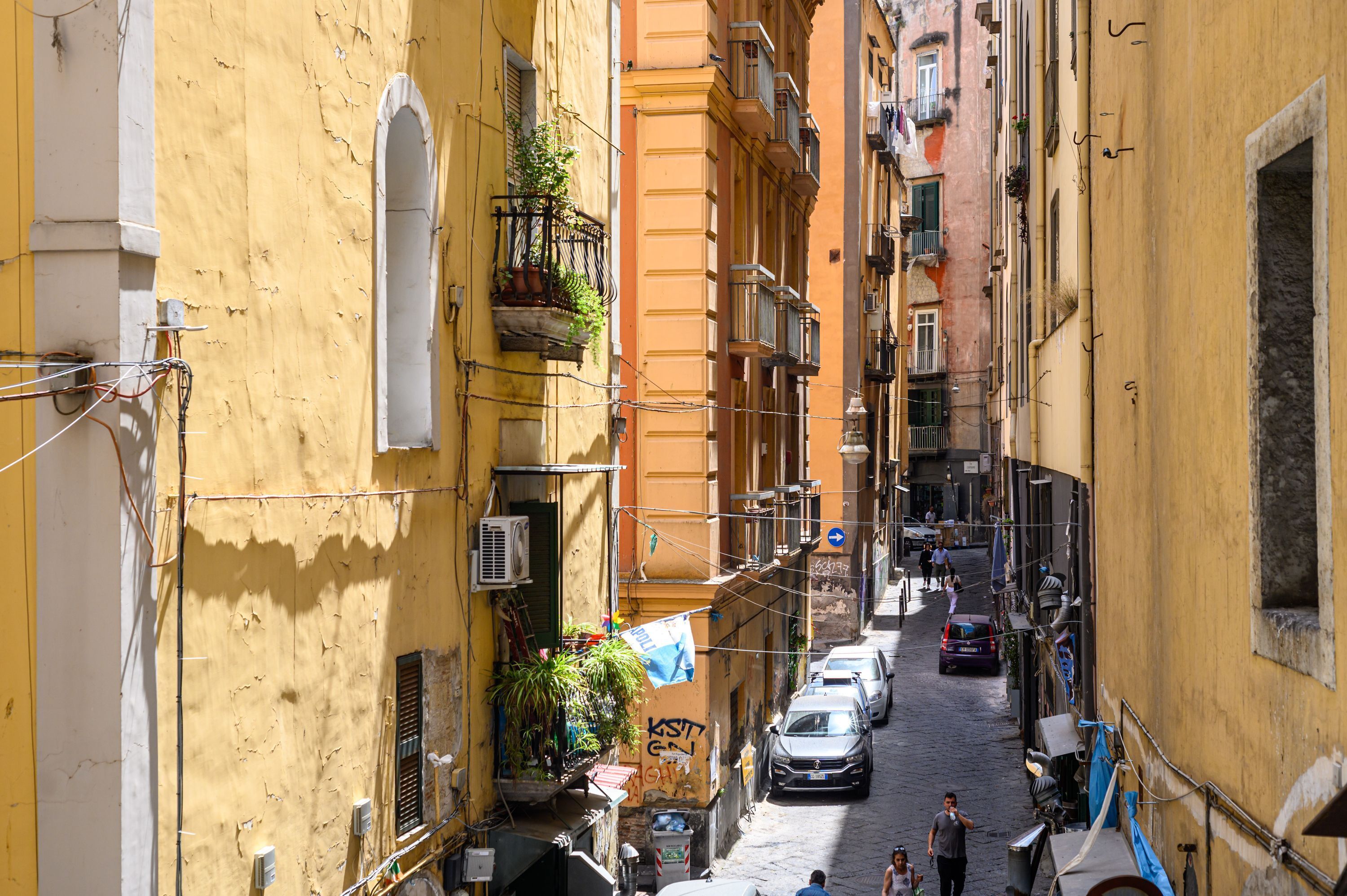 Una calle estrecha con edificios amarillos, coches aparcados y balcones con plantas.