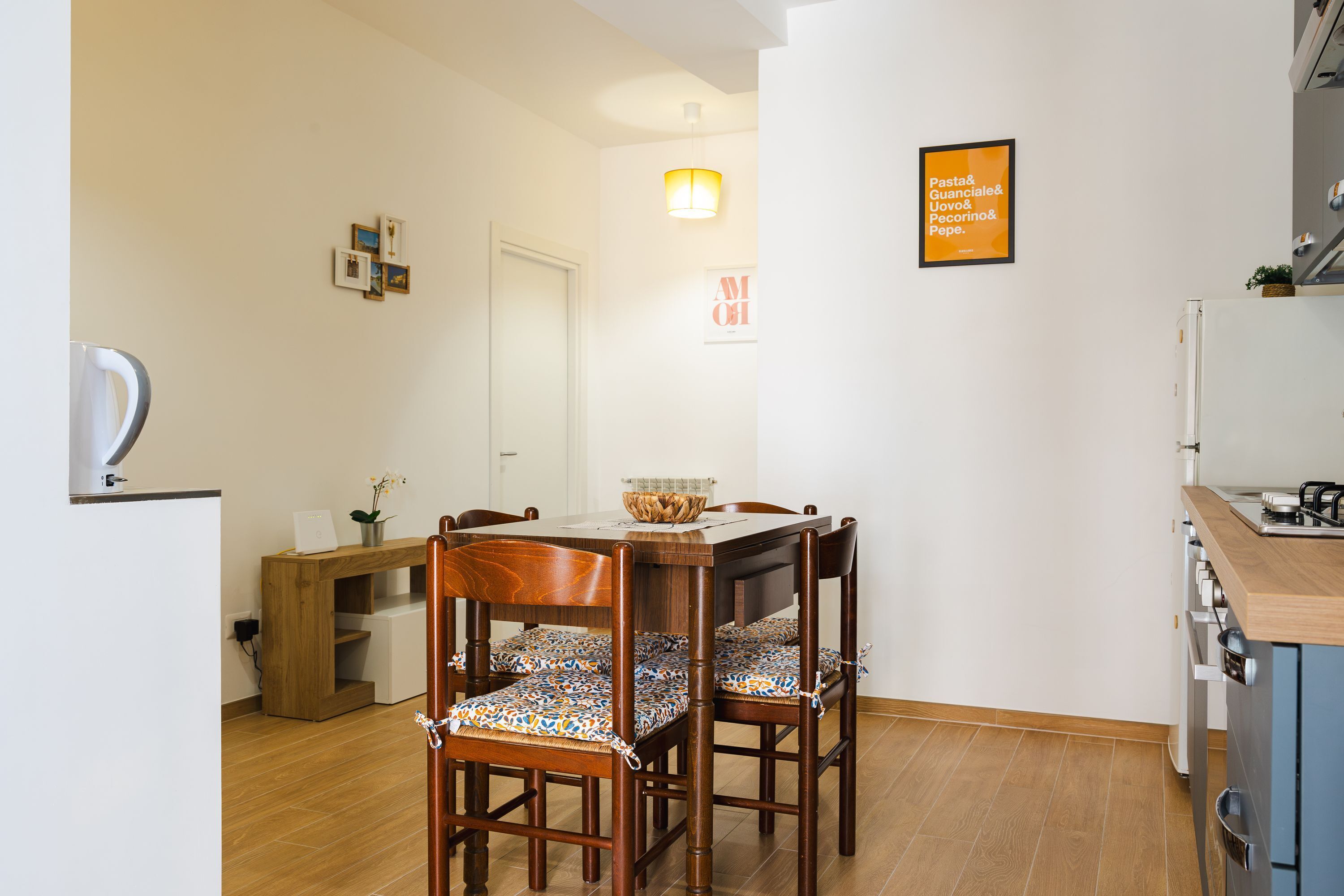 A dining area with a wooden table and chairs, a small side table with a vase, and a wall-mounted light fixture.