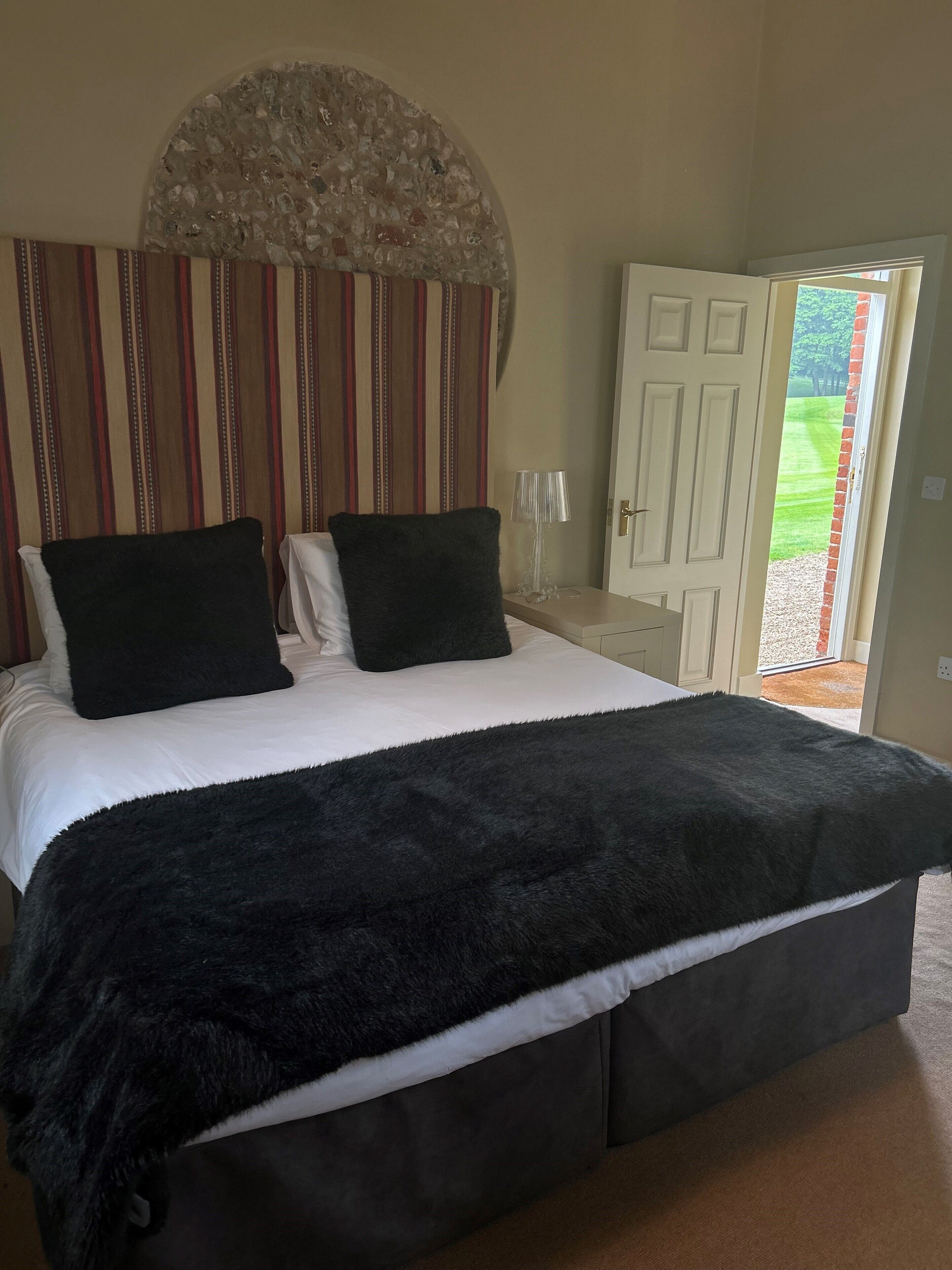 Une chambre avec un grand lit, deux oreillers noirs, une table de chevet blanche avec une lampe, une tête de lit à rayures et une vue sur un jardin par une porte ouverte.