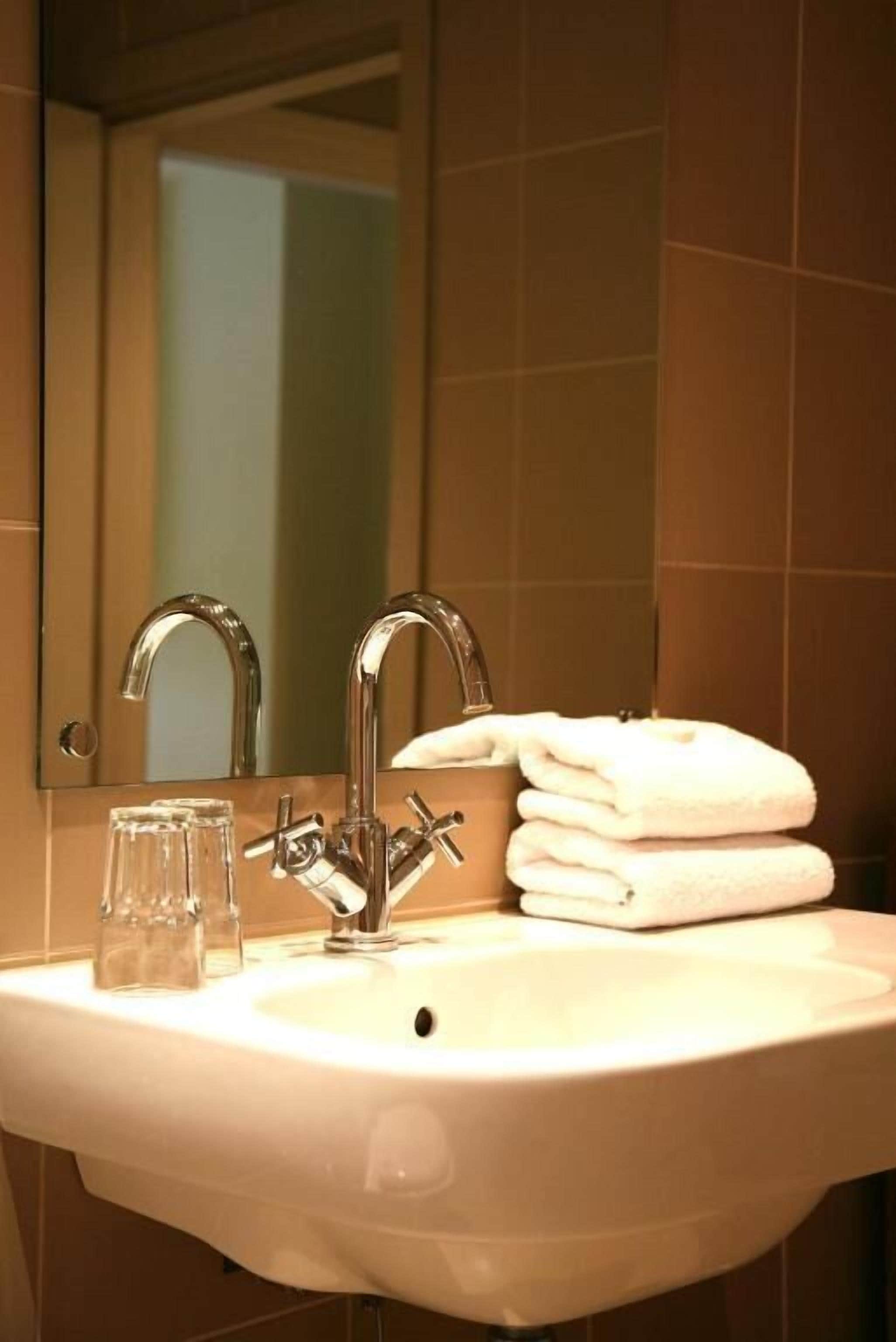 A bathroom with a sink, two faucets, and a mirror. There are two clear glasses on the sink and a stack of white towels.