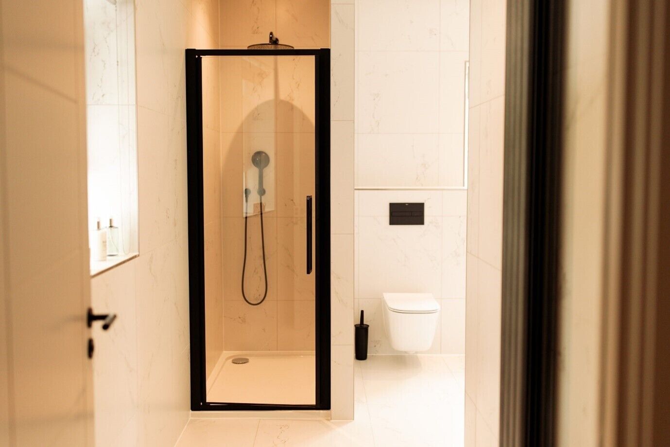 Un box doccia in vetro con soffione a mano, un water bianco e un asciugamani elettrico nero fissato a pavimento.
