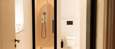 A glass-enclosed shower with a handheld showerhead, a white toilet, and a black floor-mounted hand dryer.