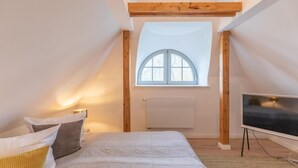 A bedroom with a sloped ceiling, a large window, a bed with pillows, a flat-screen TV, and a radiator.