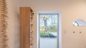 A narrow hallway with a wooden shelf unit, a white cushioned bench, and a door leading to an outdoor area with a view of trees and a fence.