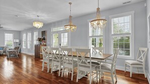 A spacious dining area with a wooden table and white chairs, large windows with blinds, and two chandeliers. - Searenity | 5BR Marshview Retreat w/ Game Room, Elevator & Close to Beach (Wrightsville Beach)