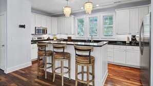 A modern kitchen with white cabinetry, a central island with bar stools, a stainless steel refrigerator, and pendant lighting. - Searenity | 5BR Marshview Retreat w/ Game Room, Elevator & Close to Beach (Wrightsville Beach)