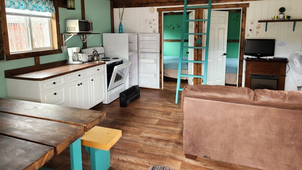 Een compacte keuken met witte kasten, een houten aanrecht en een fornuis. Een blauwgroene ladder is tegen een deur geleund, en er staat een bruine bank in de woonkamer.