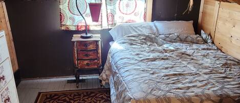A cozy attic bedroom with a sloped ceiling, a bed with patterned bedding, a small bedside table, a decorative hanging, and a window with patterned curtains.