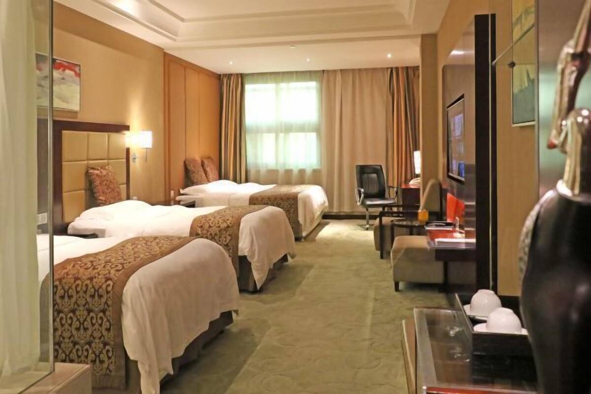 Ein Hotelzimmer mit drei Betten, einem Schreibtisch mit Computer, einem Fernseher und einer dekorativen Vase.