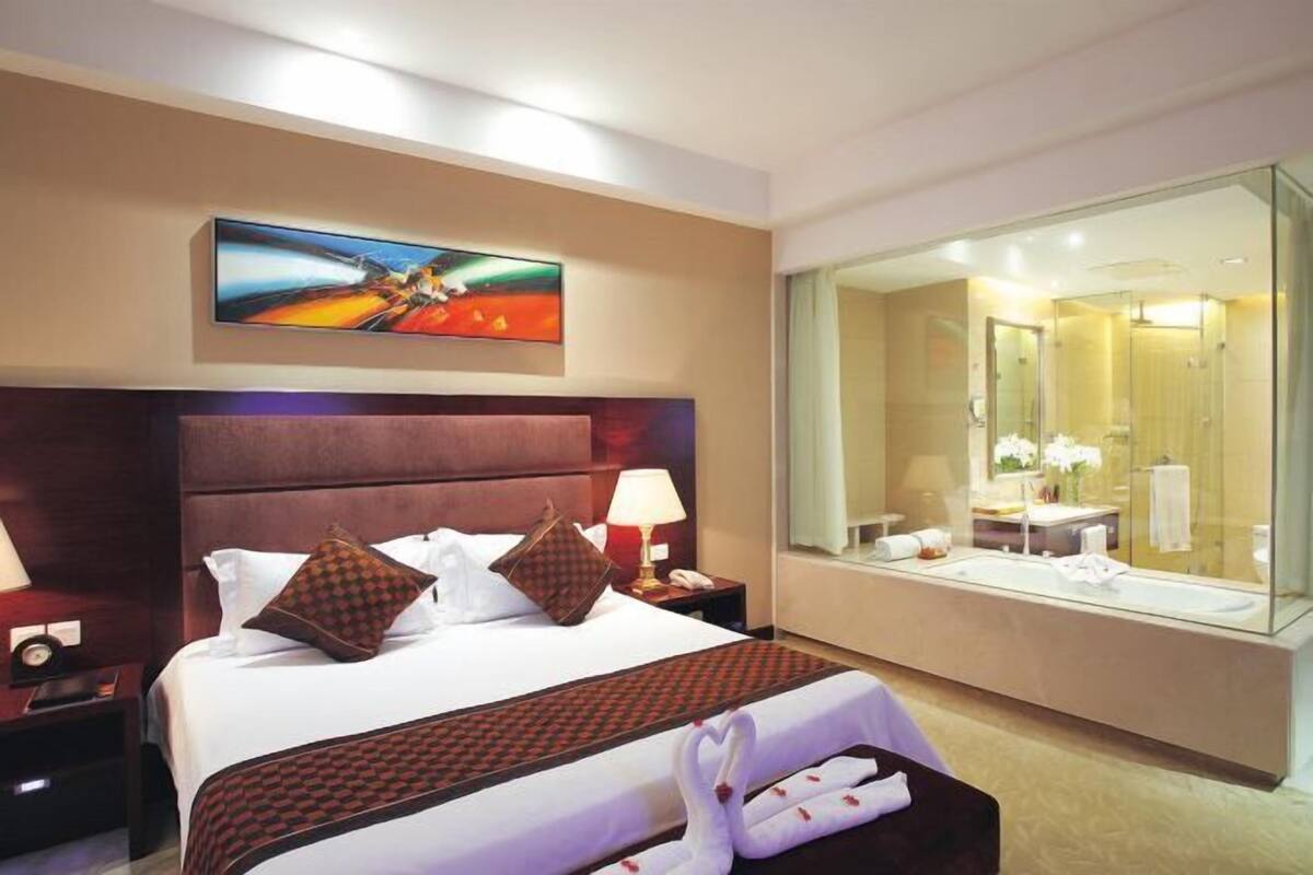 Ein Hotelzimmer mit einem großen Bett, einem Nachttisch mit Lampe, einem Gemälde an der Wand und einem großen Spiegel, der das Badezimmer reflektiert.