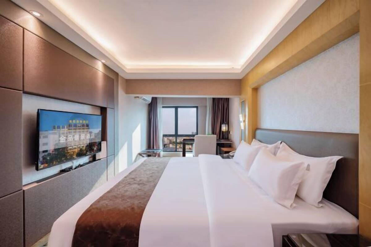 A hotel room with a large bed, a flat-screen TV mounted on the wall, a desk with a chair, and a window with curtains.