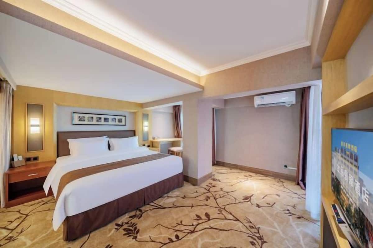 A hotel room with a large bed, a bedside table, a nightstand, a wall-mounted air conditioner, and a framed picture above the bed.